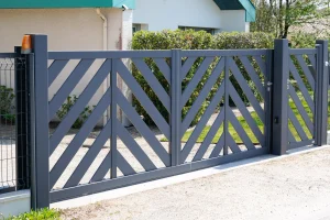 Ein modernes zweiflügeliges Gartentor aus Metall mit diagonalem Lamellendesign in Anthrazitgrau. Das Tor ist an zwei stabilen Metallpfosten befestigt, mit einem integrierten Fußgängerzugang auf der rechten Seite. Im Hintergrund befindet sich ein Haus mit einer hellen Fassade, grünen Büschen und einem gepflegten Vorgarten.