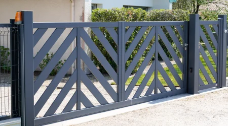 Ein modernes zweiflügeliges Gartentor aus Metall mit diagonalem Lamellendesign in Anthrazitgrau. Das Tor ist an zwei stabilen Metallpfosten befestigt, mit einem integrierten Fußgängerzugang auf der rechten Seite. Im Hintergrund befindet sich ein Haus mit einer hellen Fassade, grünen Büschen und einem gepflegten Vorgarten.