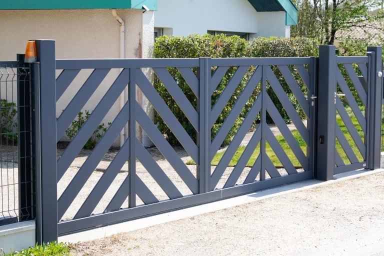 Ein modernes zweiflügeliges Gartentor aus Metall mit diagonalem Lamellendesign in Anthrazitgrau. Das Tor ist an zwei stabilen Metallpfosten befestigt, mit einem integrierten Fußgängerzugang auf der rechten Seite. Im Hintergrund befindet sich ein Haus mit einer hellen Fassade, grünen Büschen und einem gepflegten Vorgarten.