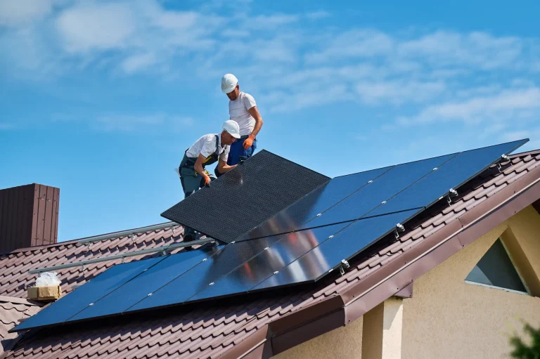 Zwei Handwerker mit Schutzkleidung montieren Photovoltaikmodule auf einem nach Süden ausgerichteten Satteldach mit braunen Dachziegeln.