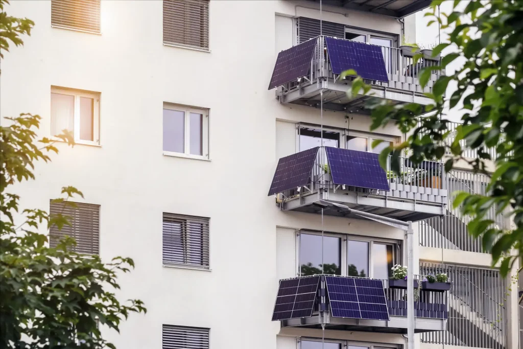 Mehrfamilienhaus mit Balkonkraftwerken – Solarpanels an mehreren Balkonen zur dezentralen Stromerzeugung, umgeben von Bäumen bei Sonnenlicht.