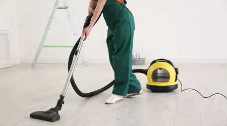 Frau in grüner Arbeitskleidung reinigt mit einem Industriestaubsauger den Boden eines frisch renovierten Raums.
