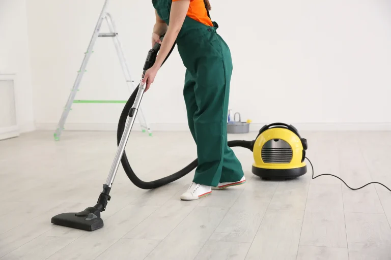 Frau in grüner Arbeitskleidung reinigt mit einem Industriestaubsauger den Boden eines frisch renovierten Raums.