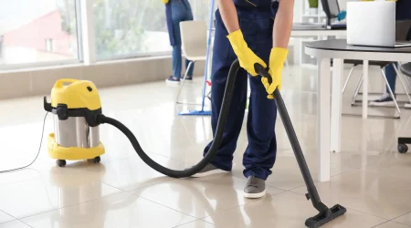 Ein Reinigungsteam säubert ein modernes Büro. Im Vordergrund saugt eine Person in blauer Arbeitskleidung und gelben Gummihandschuhen den glänzenden Fliesenboden mit einem Industriestaubsauger. Im Hintergrund reinigen zwei weitere Personen Fenster und Oberflächen von Schreibtischen.