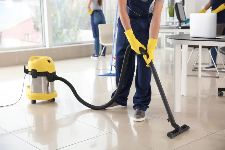 Ein Reinigungsteam säubert ein modernes Büro. Im Vordergrund saugt eine Person in blauer Arbeitskleidung und gelben Gummihandschuhen den glänzenden Fliesenboden mit einem Industriestaubsauger. Im Hintergrund reinigen zwei weitere Personen Fenster und Oberflächen von Schreibtischen.