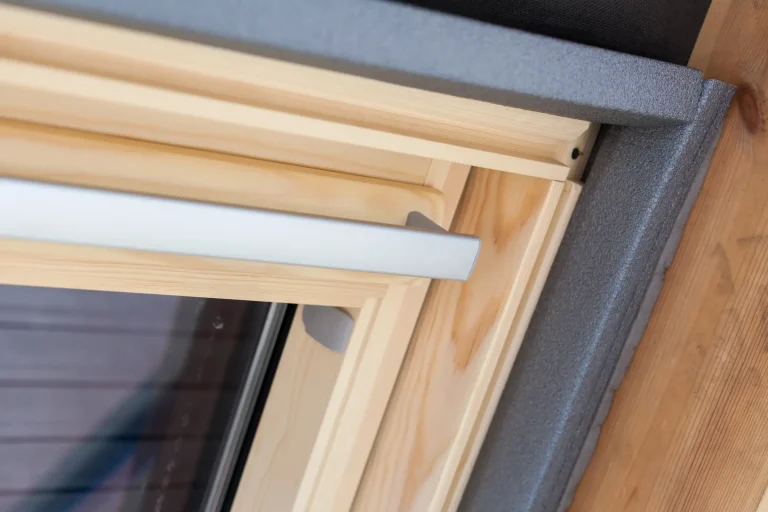 Nahaufnahme eines modernen Dachfensters mit Holzrahmen, innenliegender Isolierung und silberfarbenem Griff, fotografiert in schräger Perspektive. Die Fensterkonstruktion zeigt hochwertige Verarbeitung und Dämmmaterial zur Energieeffizienz.