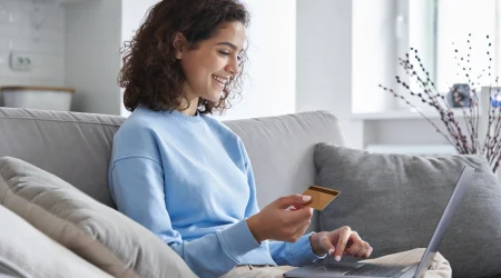 Eine Frau sitzt auf einer Couch in ihrem Wohnzimmer und schließt gerade ihr Onlinebestellung von Fenstern ab. In der Hand hält sie ihre Kreditkarte