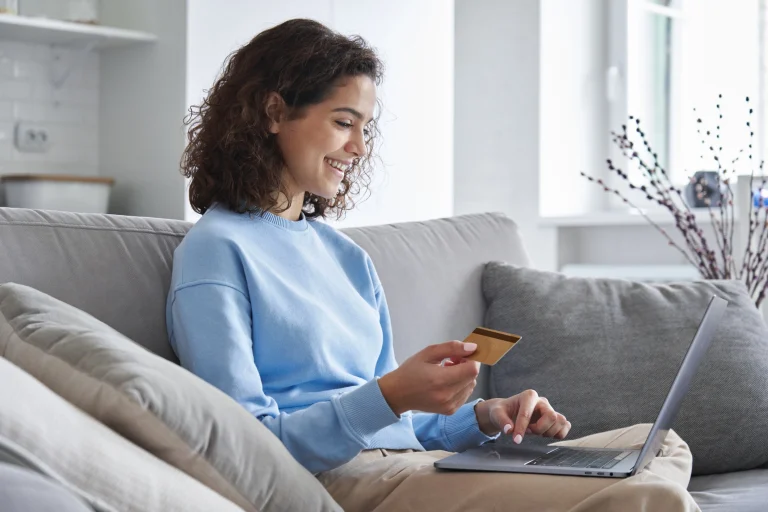 Eine Frau sitzt auf einer Couch in ihrem Wohnzimmer und schließt gerade ihr Onlinebestellung von Fenstern ab. In der Hand hält sie ihre Kreditkarte