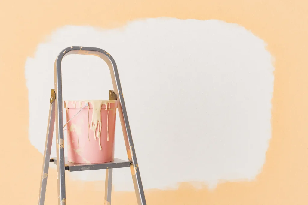 Ein Farbeimer mit flüssiger Tapete in hellem Beige steht auf einer alten, farbgesprenkelten Trittleiter vor einer halb gestrichenen Wand. Die ursprüngliche Wandfarbe ist Pfirsich-Orange, während ein großer Bereich bereits mit der beigen Flüssigtapete überstrichen wurde.