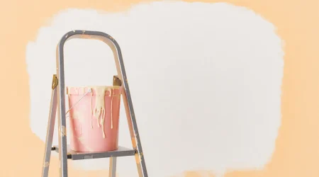 Ein Farbeimer mit flüssiger Tapete in hellem Beige steht auf einer alten, farbgesprenkelten Trittleiter vor einer halb gestrichenen Wand. Die ursprüngliche Wandfarbe ist Pfirsich-Orange, während ein großer Bereich bereits mit der beigen Flüssigtapete überstrichen wurde.