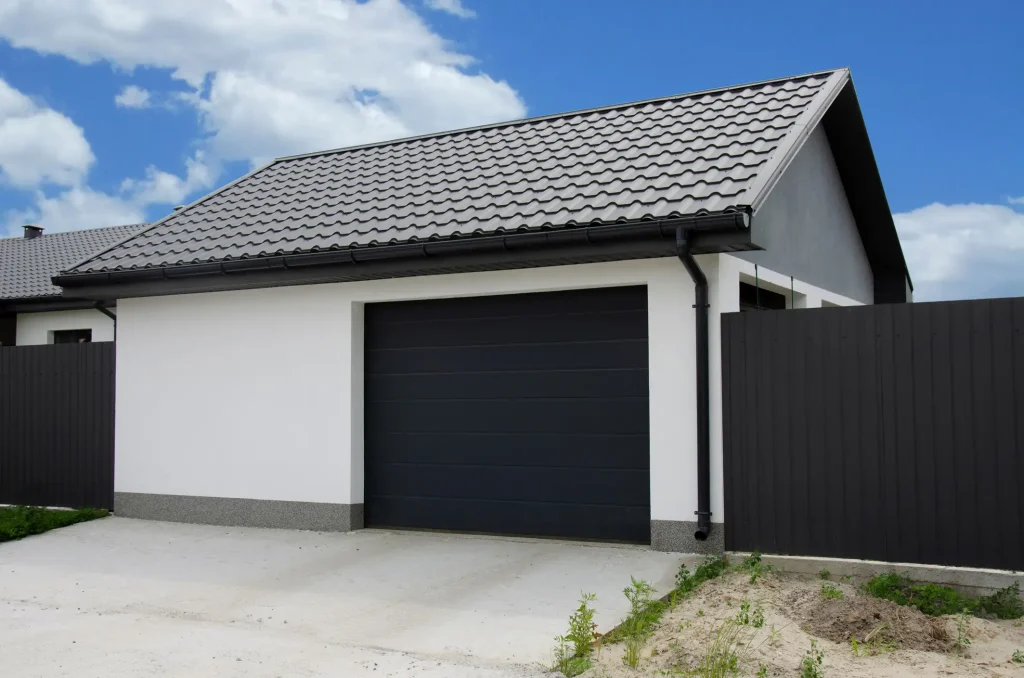 Moderne freistehende Garage mit schwarzem Sektionaltor, weißer Putzfassade und klassischem Satteldach mit anthrazitfarbener Dacheindeckung, vor blauem Himmel mit vereinzelten Wolken.