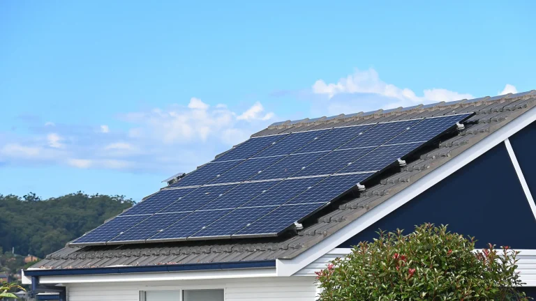 Schrägdach eines modernen Wohnhauses mit installierter Photovoltaikanlage unter blauem Himmel, im Hintergrund grüne Bäume.