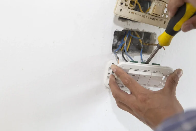 Elektriker bei der Installation eines dreifachen Lichtschalters an einer Wand – freiliegende Kabel und Unterputzdose sichtbar, Schraubendreher in der Hand.