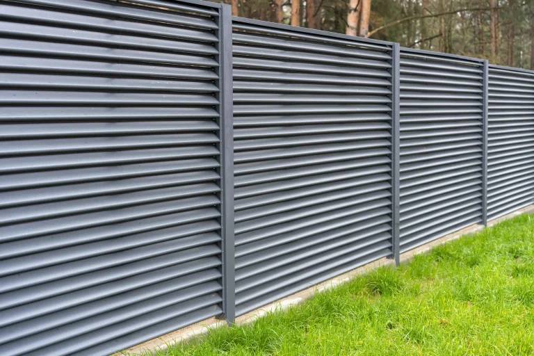 Moderne Sichtschutzzaun aus horizontalen, dunkelgrauen Metalllamellen vor einem Hintergrund aus Bäumen und grünem Rasen.