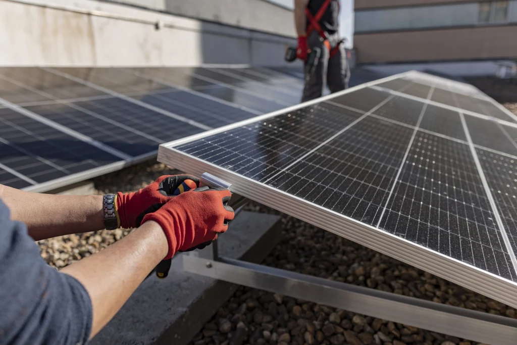 Zwei Personen montieren ein Solarmodul auf einem Flachdach. Eine Person im Vordergrund trägt rote Arbeitshandschuhe und justiert den Neigungswinkel des Moduls mithilfe eines Werkzeugs. Im Hintergrund steht eine weitere Person mit Bohrmaschine. Die Szene zeigt die präzise Ausrichtung von Photovoltaikanlagen zur optimalen Sonnenausnutzung.