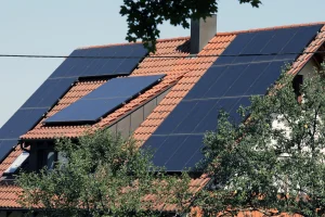 Haus mit rotem Ziegeldach, auf dem großflächig Photovoltaikmodule installiert sind. Die Solarpanels sind in verschiedenen Neigungswinkeln montiert, um den Lichteinfall optimal zu nutzen. Im Vordergrund wachsen Bäume, die teilweise die Dachfläche beschatten.
