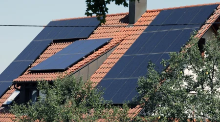 Haus mit rotem Ziegeldach, auf dem großflächig Photovoltaikmodule installiert sind. Die Solarpanels sind in verschiedenen Neigungswinkeln montiert, um den Lichteinfall optimal zu nutzen. Im Vordergrund wachsen Bäume, die teilweise die Dachfläche beschatten.