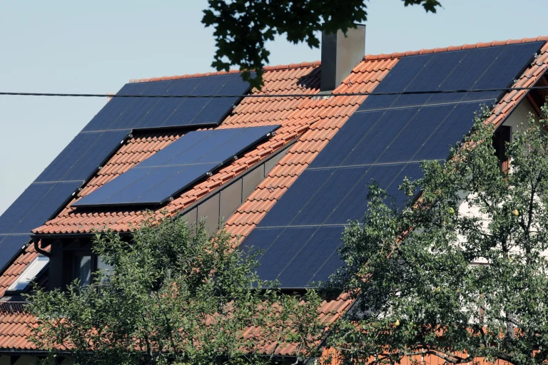 Haus mit rotem Ziegeldach, auf dem großflächig Photovoltaikmodule installiert sind. Die Solarpanels sind in verschiedenen Neigungswinkeln montiert, um den Lichteinfall optimal zu nutzen. Im Vordergrund wachsen Bäume, die teilweise die Dachfläche beschatten.