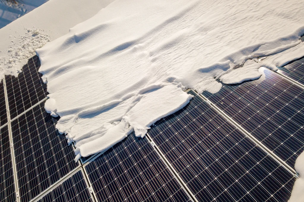 Teilweise mit Schnee bedeckte Solarmodule auf einem Dach. Der Schnee beginnt, durch den Neigungswinkel der Module nach unten zu rutschen, wodurch ein Teil der Photovoltaikfläche wieder freiliegt. Das Bild verdeutlicht die Bedeutung des Neigungswinkels für die Schneeräumung und Effizienz von Solaranlagen im Winter.