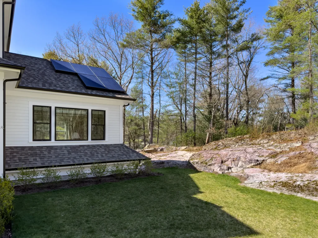 Teilansicht eines modernen Hauses mit Satteldach mit Photovoltaikmodulen, auf denen teilweise Schatten durch den Dachüberstand fällt, umgeben von Garten, Felsboden und Bäumen an einem sonnigen Tag.