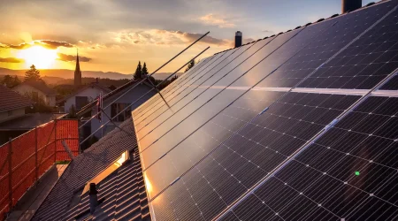 Photovoltaikanlage auf einem Hausdach bei Sonnenuntergang, mit Blick auf benachbarte Häuser, Bäume und eine Kirche im Hintergrund.