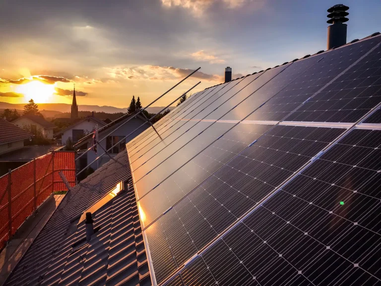 Photovoltaikanlage auf einem Hausdach bei Sonnenuntergang, mit Blick auf benachbarte Häuser, Bäume und eine Kirche im Hintergrund.
