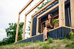 Frau sitzt mit ihrem Smartphone auf der kleinen Terrasse ihres Tiny Houses
