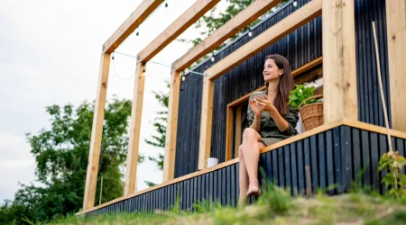 Frau sitzt mit ihrem Smartphone auf der kleinen Terrasse ihres Tiny Houses