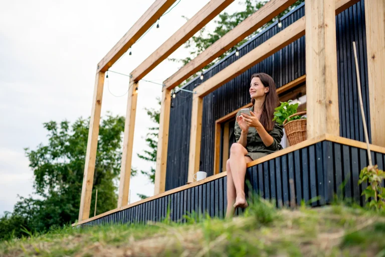 Frau sitzt mit ihrem Smartphone auf der kleinen Terrasse ihres Tiny Houses