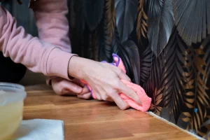 Person reinigt mit einem Mikrofasertuch den unteren Rand einer abwaschbaren Vinyltapete an der Wand; sichtbar strukturierte Oberfläche mit dunklem, tropischem Muster auf Holzboden.