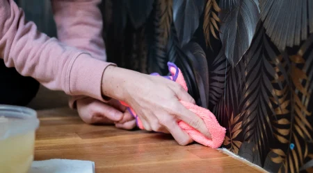 Person reinigt mit einem Mikrofasertuch den unteren Rand einer abwaschbaren Vinyltapete an der Wand; sichtbar strukturierte Oberfläche mit dunklem, tropischem Muster auf Holzboden.