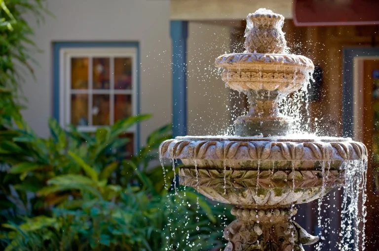 Nahaufnahme eines kunstvoll gestalteten Steinbrunnens mit fließendem Wasser in einem grünen Garten, im Hintergrund Pflanzen und ein Fenster mit blauer Umrahmung.