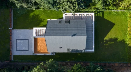 Modernes Wohnhaus mit dunkel gedecktem Satteldach in symmetrischer Form, aus der Vogelperspektive aufgenommen. Das Dach verläuft gleichmäßig in zwei geneigten Flächen von einem zentralen First abwärts. Seitlich befinden sich zwei Dachfenster, das Gebäude ist von einem gepflegten Garten mit Rasen und gepflasterten Terrassen umgeben.