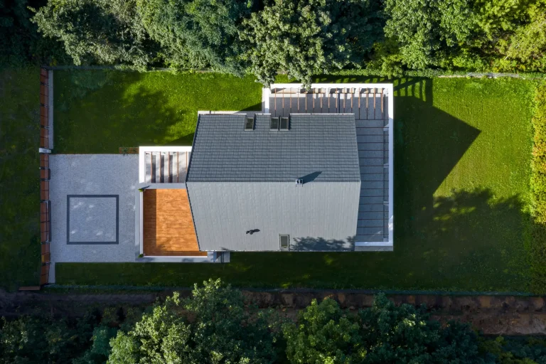 Modernes Wohnhaus mit dunkel gedecktem Satteldach in symmetrischer Form, aus der Vogelperspektive aufgenommen. Das Dach verläuft gleichmäßig in zwei geneigten Flächen von einem zentralen First abwärts. Seitlich befinden sich zwei Dachfenster, das Gebäude ist von einem gepflegten Garten mit Rasen und gepflasterten Terrassen umgeben.