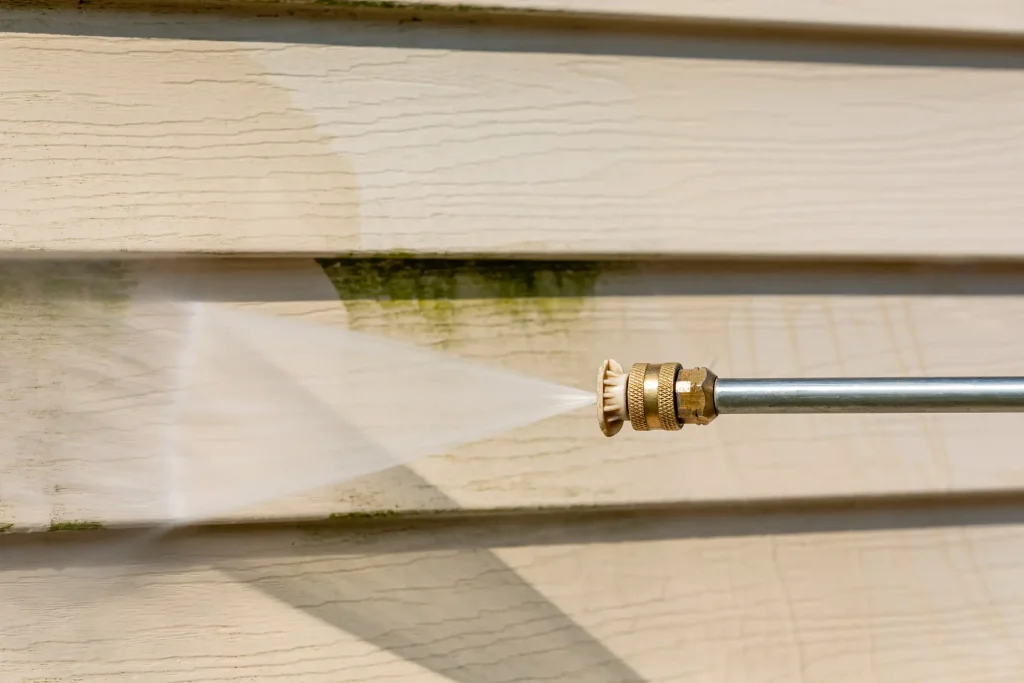 Nahaufnahme eines Hochdruckreinigers bei der Reinigung einer Hausfassade mit Kunststoffverkleidung; deutlich sichtbare Entfernung von grünem Algenbefall durch den Wasserstrahl.