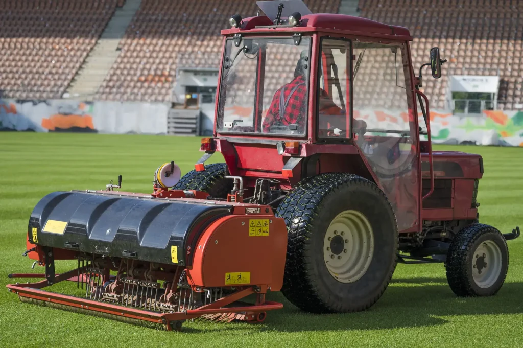 Traktor mit angebautem Rasenbelüfter bei der mechanischen Aerifizierung eines Fußball- oder Sportplatzes, zur Verbesserung der Bodenbelüftung und Drainage.