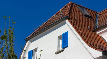 Weiß verputztes Wohnhaus mit leuchtend blauen Fensterläden und einem rot gedeckten Krüppelwalmdach. Das Dach ist an der Giebelseite nicht vollständig abgewalmt – der obere Teil bildet ein typisches Satteldach, während der untere Bereich abgeschrägt ist. Eine kleine Gaube sowie Schneefanggitter und Blitzschutzleitungen ergänzen die Dachkonstruktion.