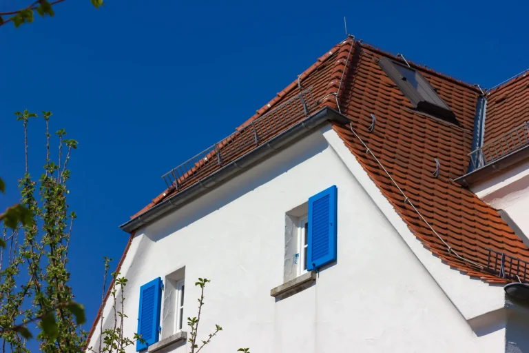 Weiß verputztes Wohnhaus mit leuchtend blauen Fensterläden und einem rot gedeckten Krüppelwalmdach. Das Dach ist an der Giebelseite nicht vollständig abgewalmt – der obere Teil bildet ein typisches Satteldach, während der untere Bereich abgeschrägt ist. Eine kleine Gaube sowie Schneefanggitter und Blitzschutzleitungen ergänzen die Dachkonstruktion.