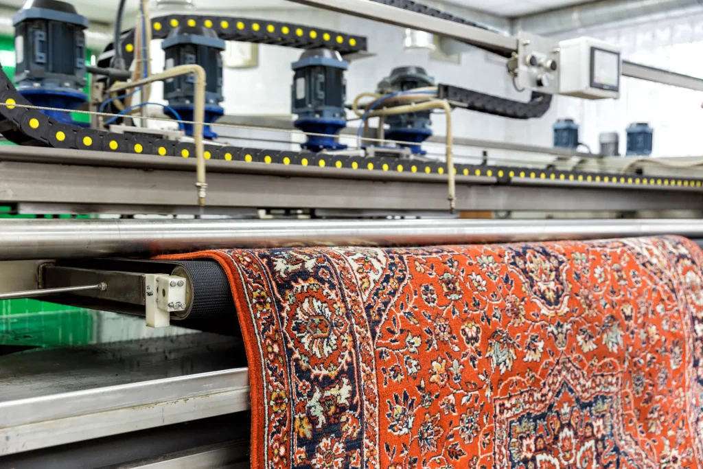 Teppich mit orientalischem Muster wird in einer industriellen Reinigungsmaschine maschinell gesäubert – Nahaufnahme einer rot-blauen Teppichkante im Waschvorgang.