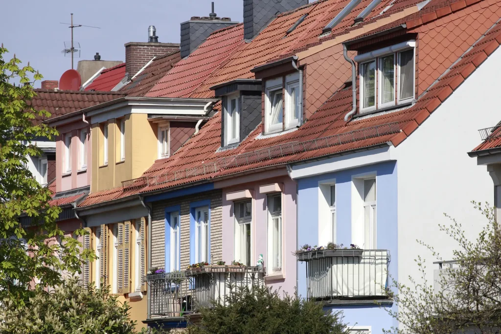 Reihenhäuser mit einzelnen Satteldächern, die nahtlos aneinander anschließen und optisch wie ein durchgehendes Dach wirken. Die Dächer sind mit roten Ziegeln gedeckt, einige besitzen Gauben und Dachfenster. Die bunt gestrichenen Fassaden unterstreichen die architektonische Gliederung der einzelnen Häuser.