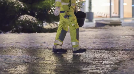 Winterdienstmitarbeiter in reflektierender Arbeitskleidung geht mit einem Eimer über einen glatten Gehweg vor einem Gebäude bei Schneefall.