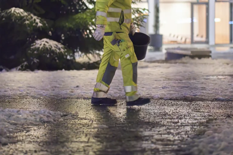 Winterdienstmitarbeiter in reflektierender Arbeitskleidung geht mit einem Eimer über einen glatten Gehweg vor einem Gebäude bei Schneefall.