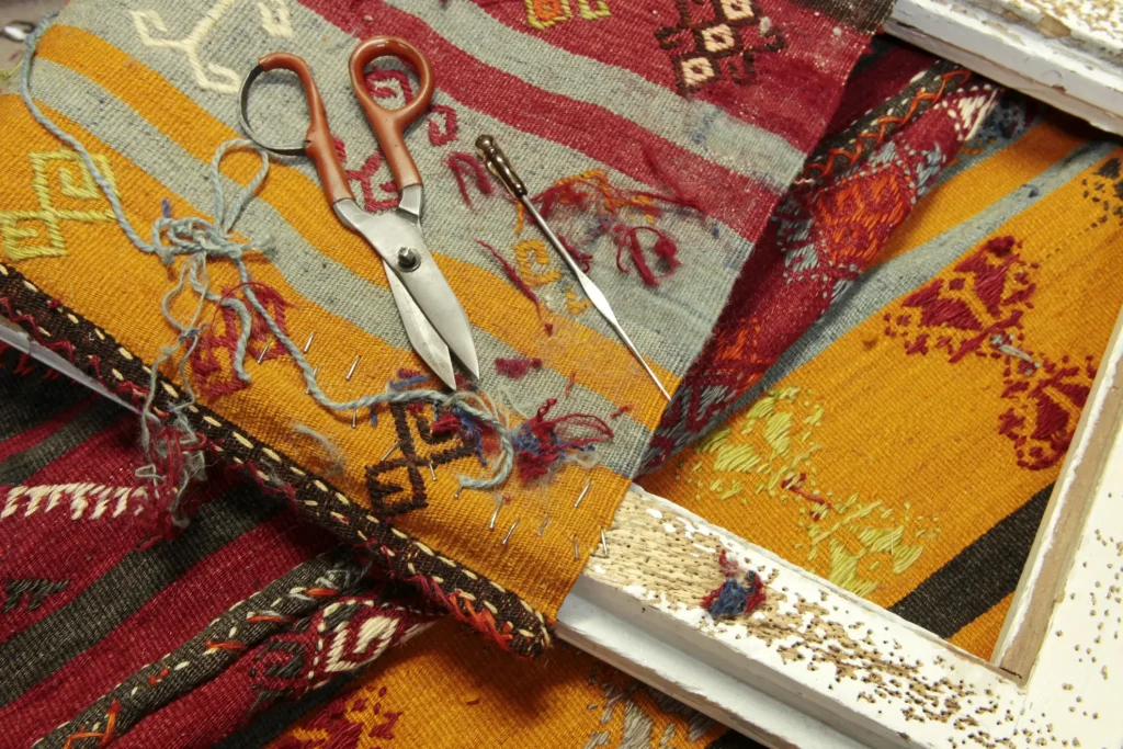 Detailaufnahme eines bunten, handgewebten Teppichs in Reparatur mit Schere, Nadel und Werkzeug auf der Oberfläche.