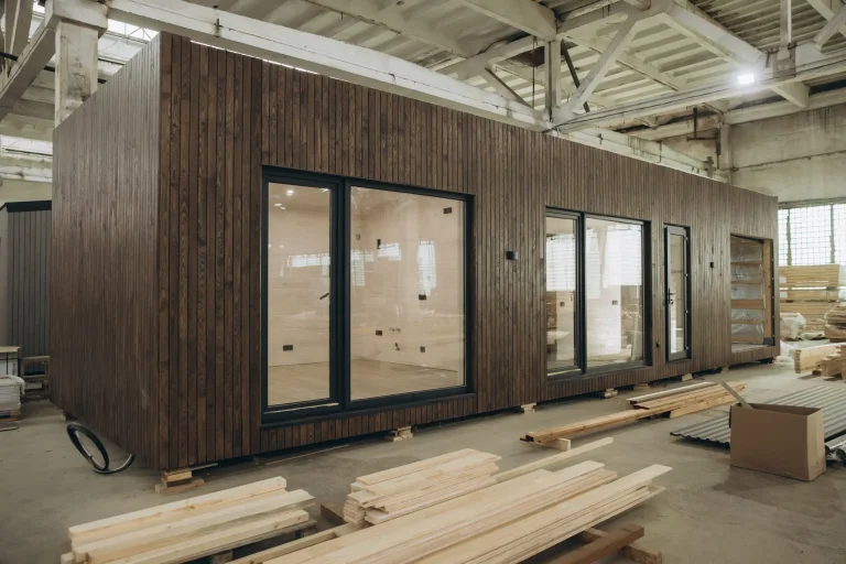 Modernes Tiny House mit dunkler Holzfassade und großen Glasfronten in einer Produktionshalle; im Vordergrund liegen Holzbretter für den Innenausbau.