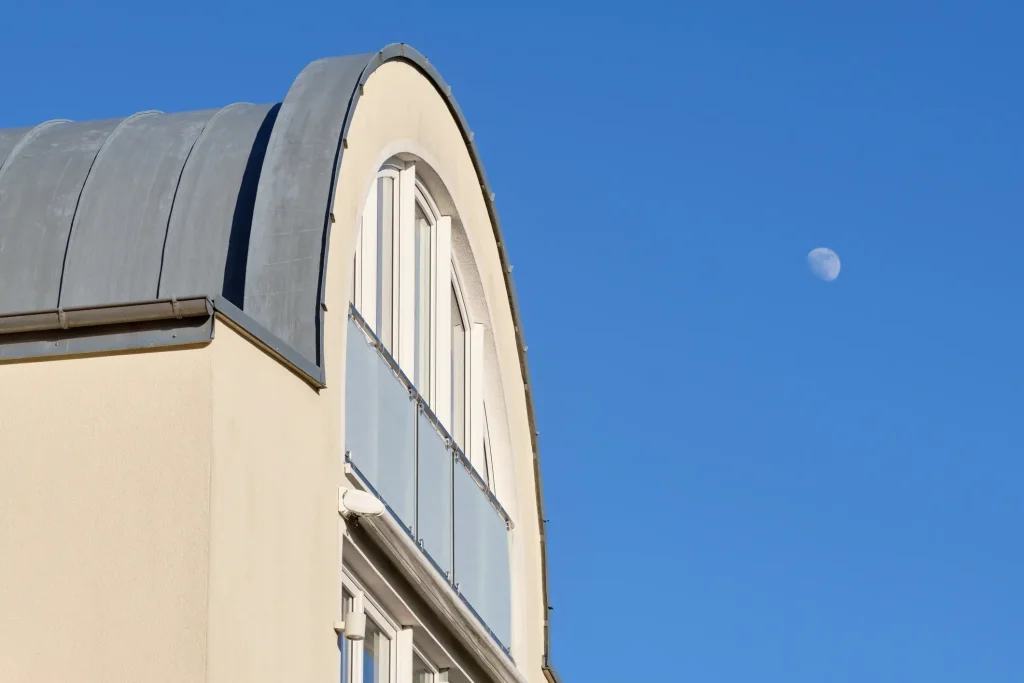 Modernes Wohngebäude mit hellem Putz und einem abgerundeten Tonnendach aus Metall. Am Himmel ist der Mond zu sehen.
