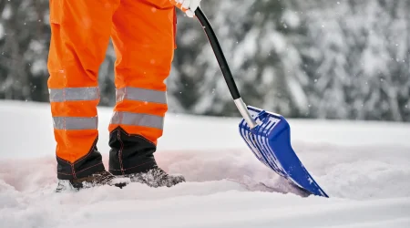 Eine Person im Winterdienst schiebt Schnee mit einer Schneeschaufel auf einem verschneiten Gehweg.