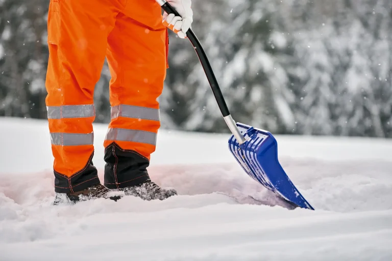 Eine Person im Winterdienst schiebt Schnee mit einer Schneeschaufel auf einem verschneiten Gehweg.