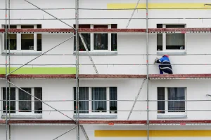 Ein Handwerker steht auf einem Baugerüst vor einer Wohnhausfassade und bringt Aerogel-Dämmplatten an. Das Bild zeigt farbige Fassadenbereiche und mehrere Fenster.