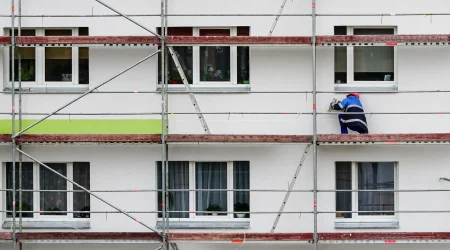 Ein Handwerker steht auf einem Baugerüst vor einer Wohnhausfassade und bringt Aerogel-Dämmplatten an. Das Bild zeigt farbige Fassadenbereiche und mehrere Fenster.