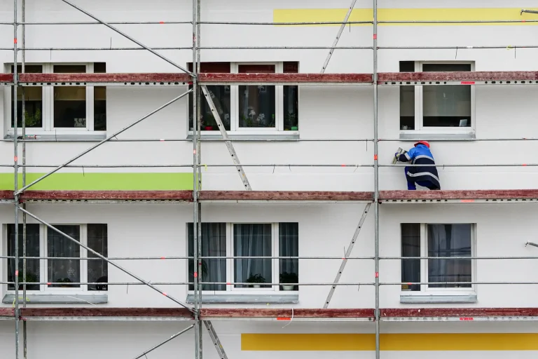 Ein Handwerker steht auf einem Baugerüst vor einer Wohnhausfassade und bringt Aerogel-Dämmplatten an. Das Bild zeigt farbige Fassadenbereiche und mehrere Fenster.